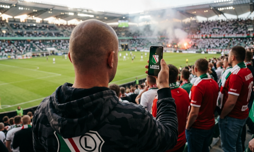 Białoskóry łysy kibic Legii Warszawa trzyma smartfon z logo Legia Mobile. Ujęcie zza pleców promujące ofertę komórkową dla kibiców w 2026 roku.