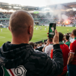 Białoskóry łysy kibic Legii Warszawa trzyma smartfon z logo Legia Mobile. Ujęcie zza pleców promujące ofertę komórkową dla kibiców w 2026 roku.