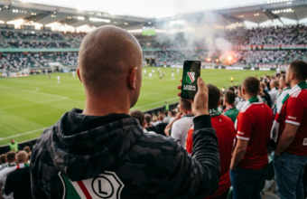 Białoskóry łysy kibic Legii Warszawa trzyma smartfon z logo Legia Mobile. Ujęcie zza pleców promujące ofertę komórkową dla kibiców w 2026 roku.