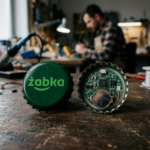 Zielony kapsel z logo Żabka i modułem elektronicznym. Kontroler Żappka Triki to gadżet gamingowy i element kultury cyfrowej polskiej sieci sklepów.