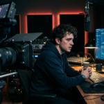 Zestresowany twórca YouTube w studio analizuje wykresy. Monetyzacja YouTube (Polska i świat) martwi rynek tech. Niebieskie światło monitora i rozmyta kamera.