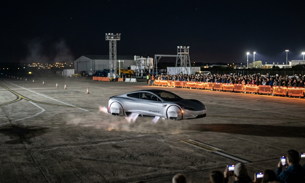 Nowa srebrna futurystyczna Tesla Roadster z silnikami SpaceX unosi się nocą dzięki wywietrznikom w podwoziu i bokach. Widzowie w oddali na torze SpaceX.